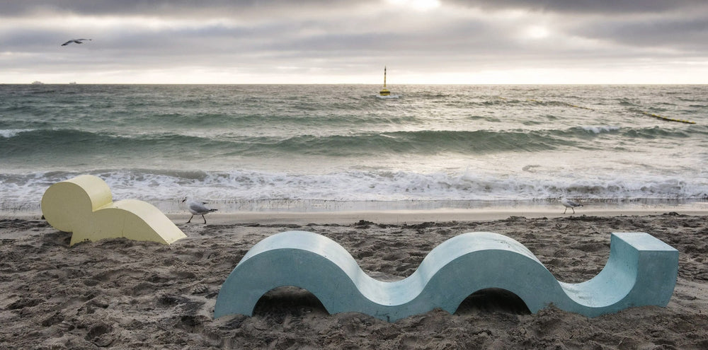 Two abstract concrete sculptures, one a curved yellow figure suggesting a swimmer’s body, the other a pale blue wave form, positioned on the sand at Cottesloe Beach, Western Australia.