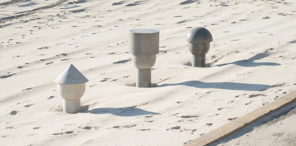 Three abstract concrete heads — one white, one mid-grey, one dark grey — partially embedded in the sand at Cottesloe Beach, Western Australia.