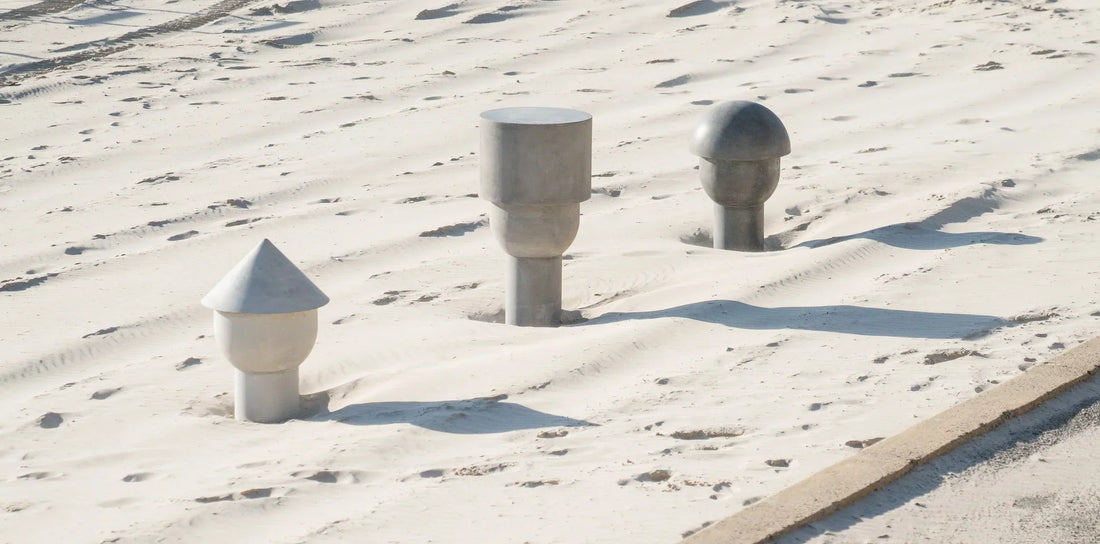 Three abstract concrete heads — one white, one mid-grey, one dark grey — partially embedded in the sand at Cottesloe Beach, Western Australia.