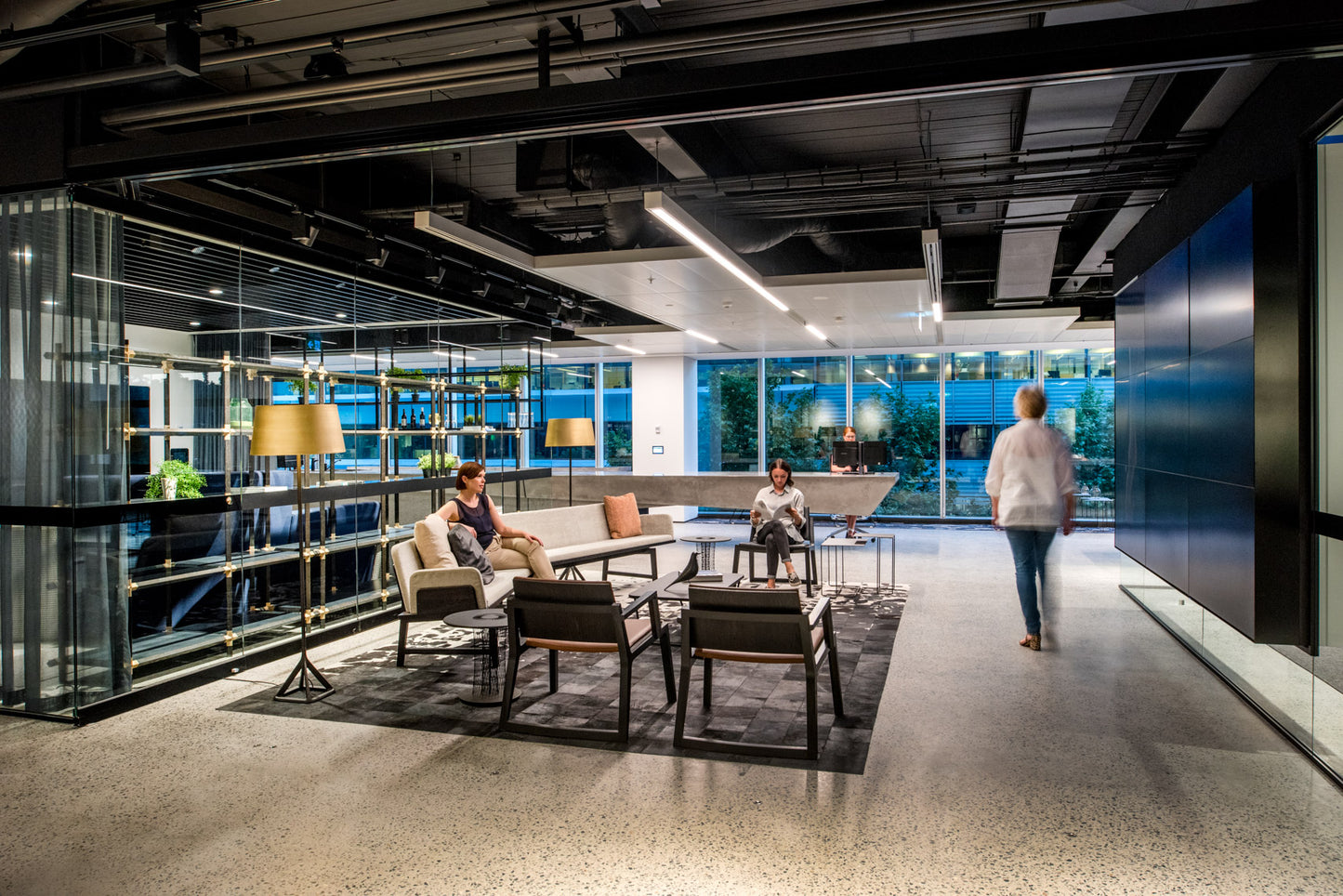 Modern office lobby with people sitting on a couch. One person is walking towards a large concrete reception desk.