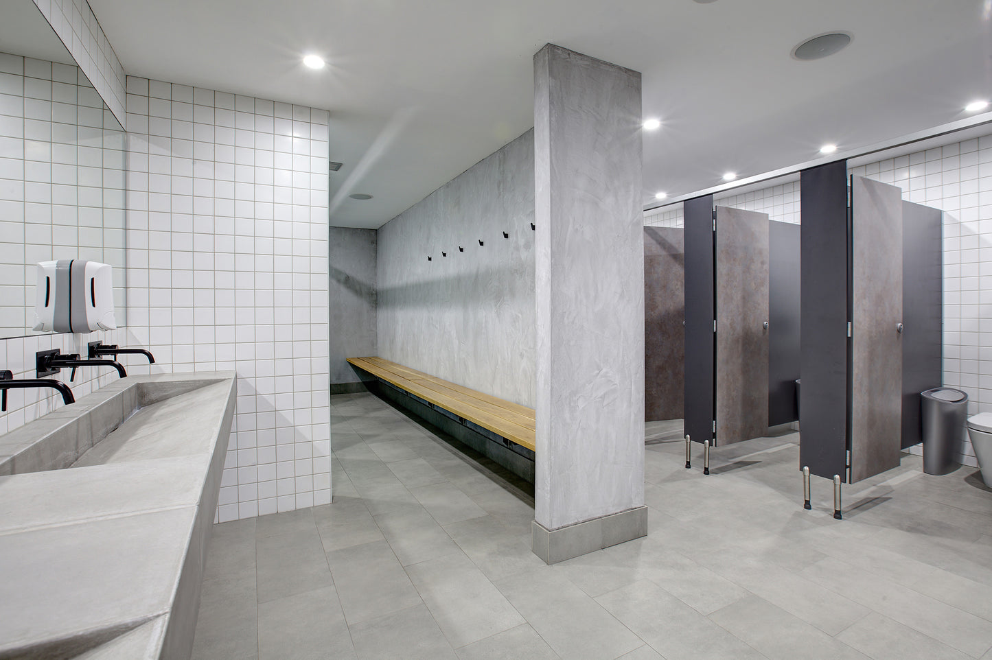 Modern bathroom with concrete ramp sinks, tiled walls, a long bench, and stalls.