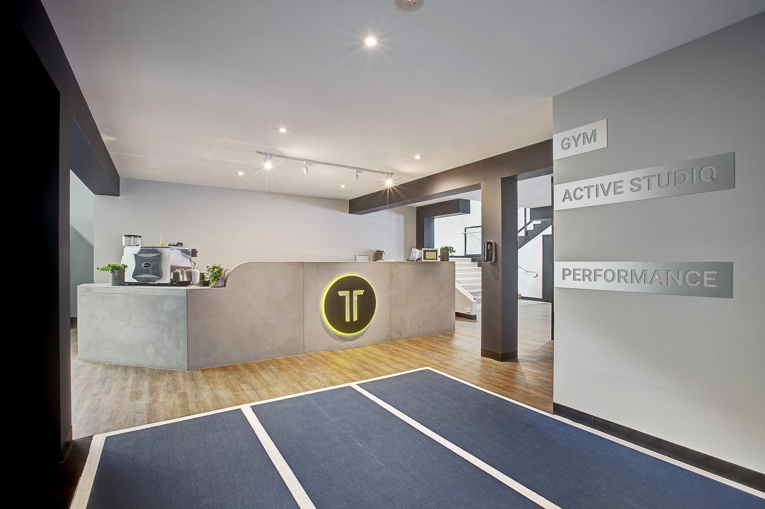 Modern interior of a gym with a concrete architectural reception desk and 'GYM ACTIVE STUDIO PERFORMANCE' signs on the walls.