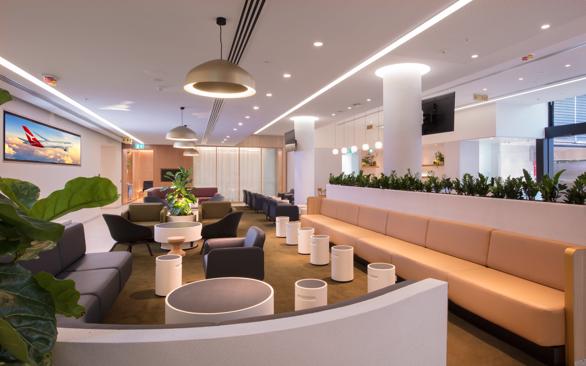 Modern airport business class lounge with concrete cylindrical tables, seating area, plants, and a TV screen.